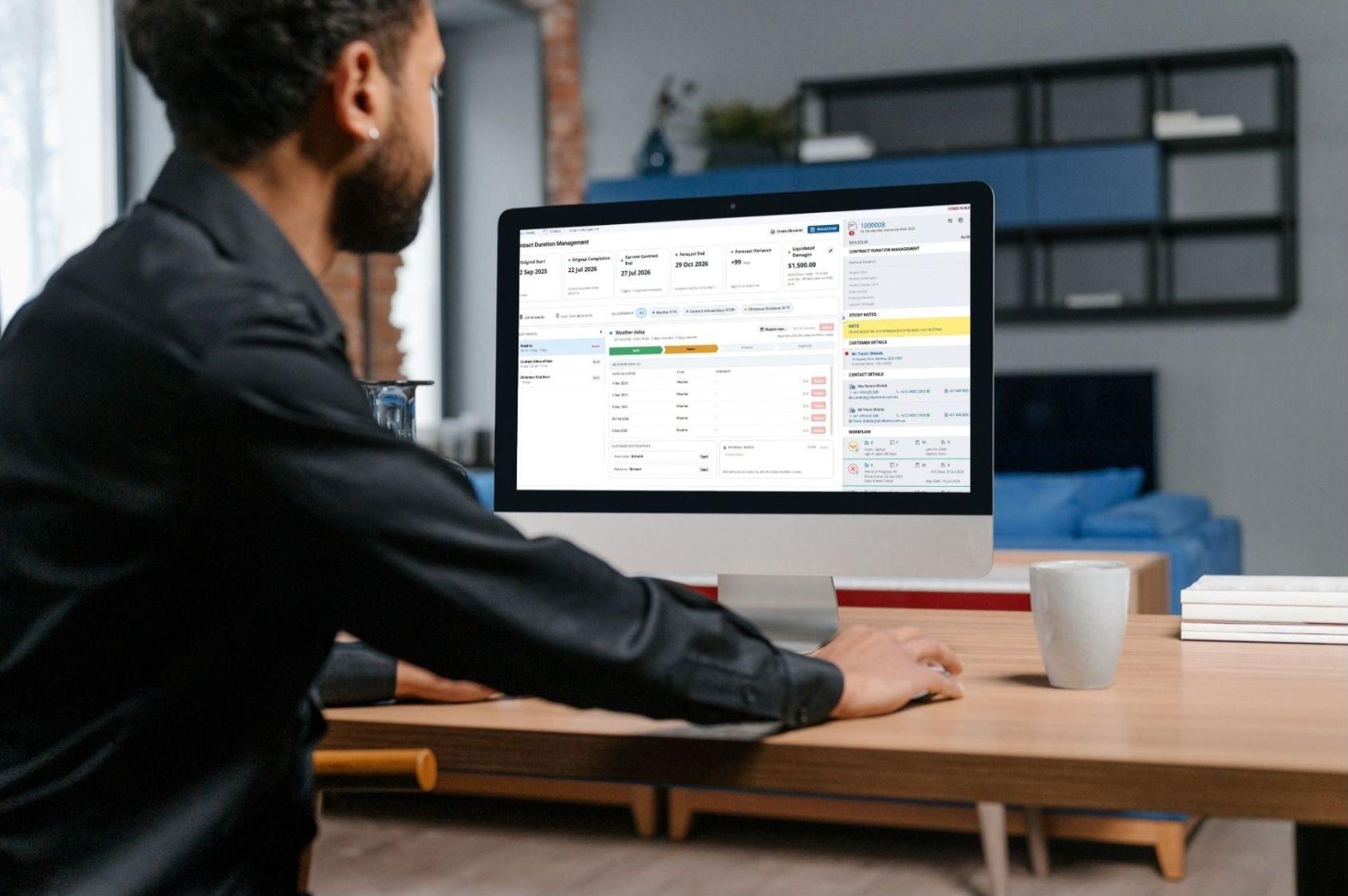 A builder at a desktop computer reviewing the ClickHome27 Contract Duration Management screen, showing contract dates, forecast variance and liquidated damages