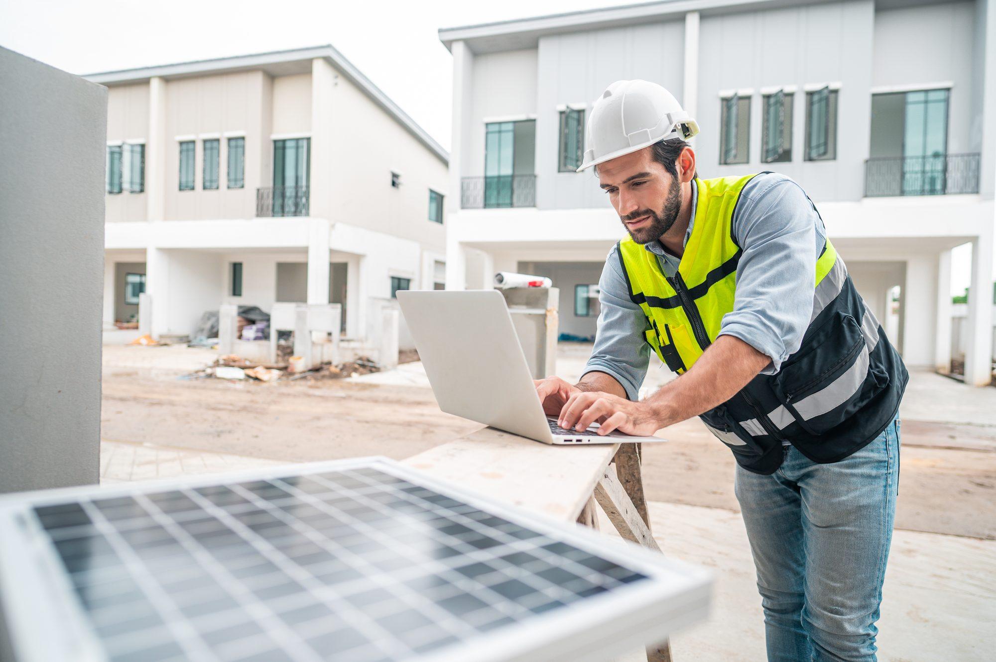 A site supervisor in a hard hat and hi-vis vest working on a laptop at a residential construction site