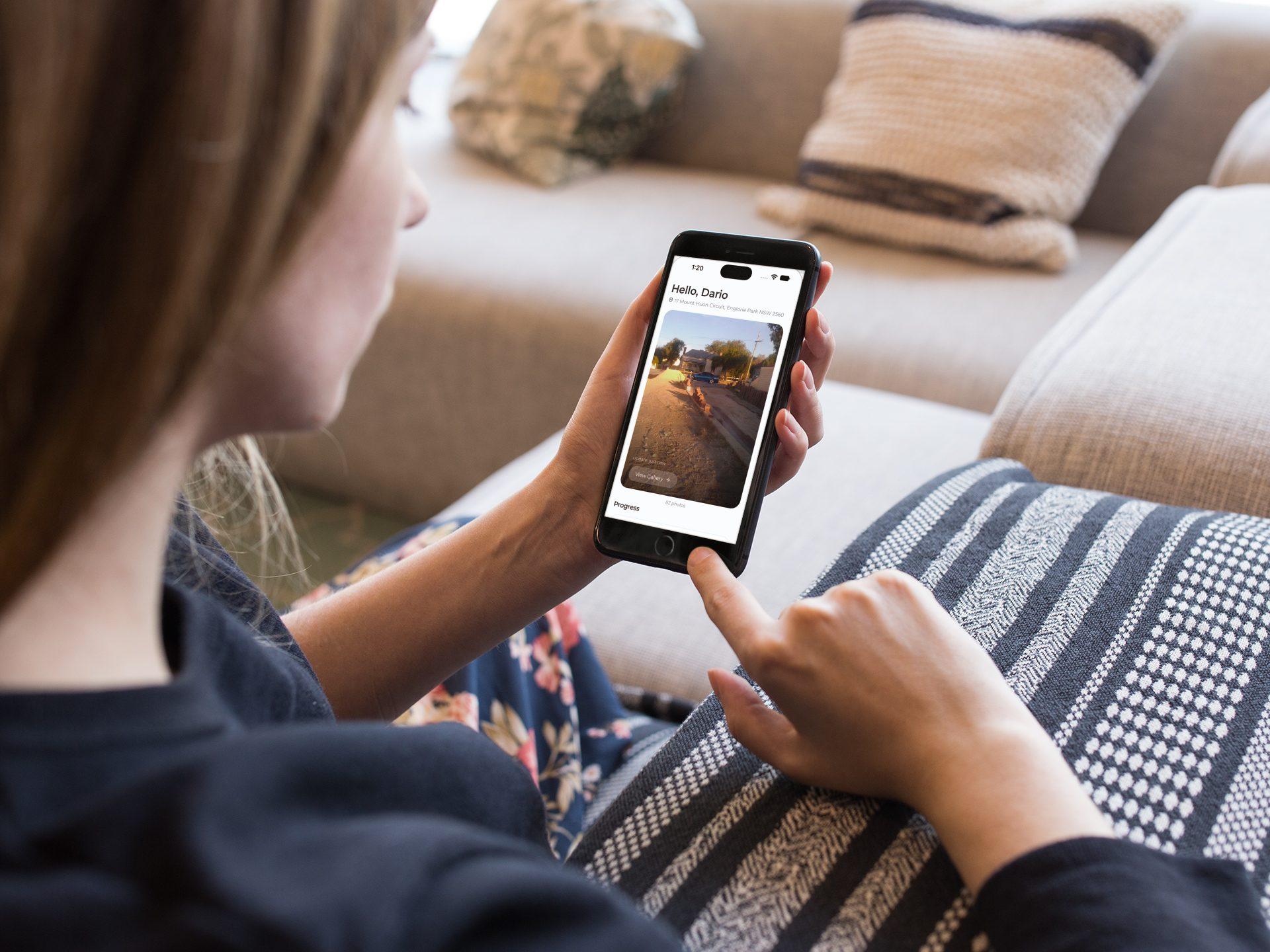 A homeowner on a sofa checking build progress photos in the MyHome mobile app on her phone